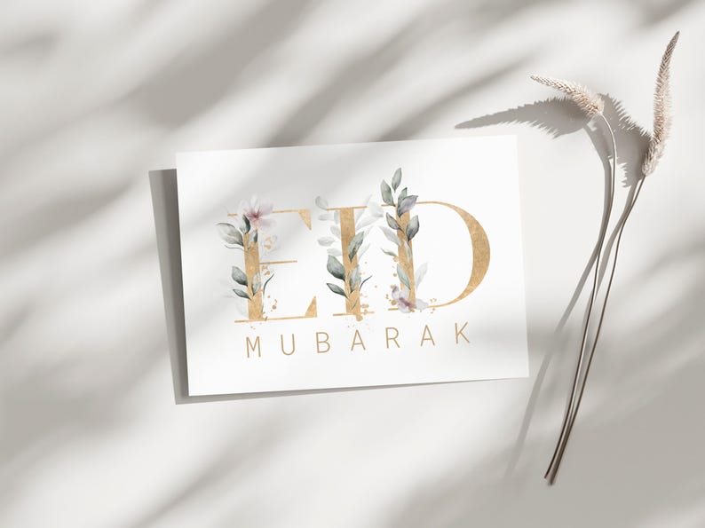May include: A white card with gold lettering that reads "Eid Mubarak". The letters are decorated with watercolor floral designs in shades of green and pink.