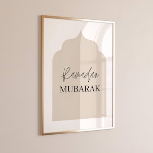 Puede incluir: Una impresión artística enmarcada con un marco dorado. La impresión presenta un diseño de arco beige con las palabras "Ramadan MUBARAK" en escritura negra y letras de bloque. El fondo es de color beige claro.