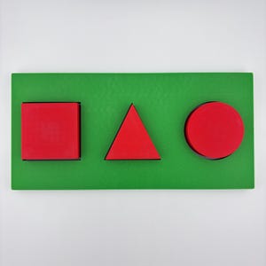 Peut inclure: Un tableau en bois vert avec trois formes géométriques rouges : un carré, un triangle et un cercle. Les formes sont en relief et ont des contours noirs. Le fond est de couleur verte unie.