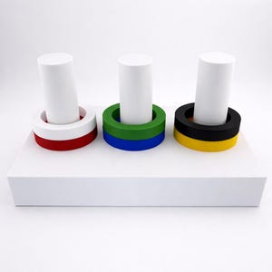May include: Three white cylindrical objects are each set in a colorful ring base. The rings are red and white, green and blue, and black and yellow. The objects sit on a white rectangular platform against a white background.