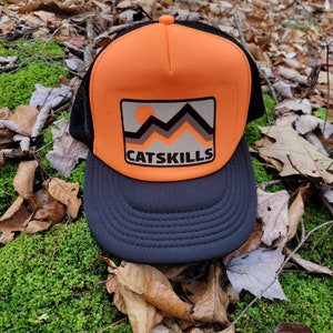 Puede incluir: Una gorra de camionero negra y naranja con un parche que dice "CATSKILLS" y un gráfico de montañas y un sol.