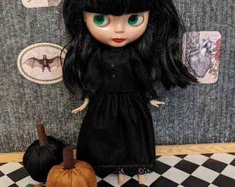 May include: A black-haired doll wearing a black dress with lace trim stands on a black and white checkered floor.  The doll has green eyes and red lips.  There are four small orange pumpkins and one black pumpkin on the floor.