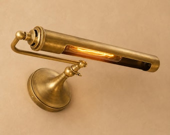 Op de afbeelding: Een wandlamp voor schilderijen met een antieke messing afwerking. Het armatuur heeft een cilindrische, horizontale lampkop met een open gleuf die een gloeiende gloeidraadlamp onthult, verbonden met een ronde voet door een verstelbare arm.