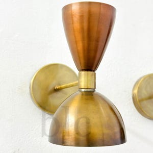 May include: A pair of wall sconces with a mid-century modern design. The sconces feature a copper-colored conical shade and a brass-colored, dome-shaped base. The sconces are mounted on a white wall with brass-colored circular backplates.