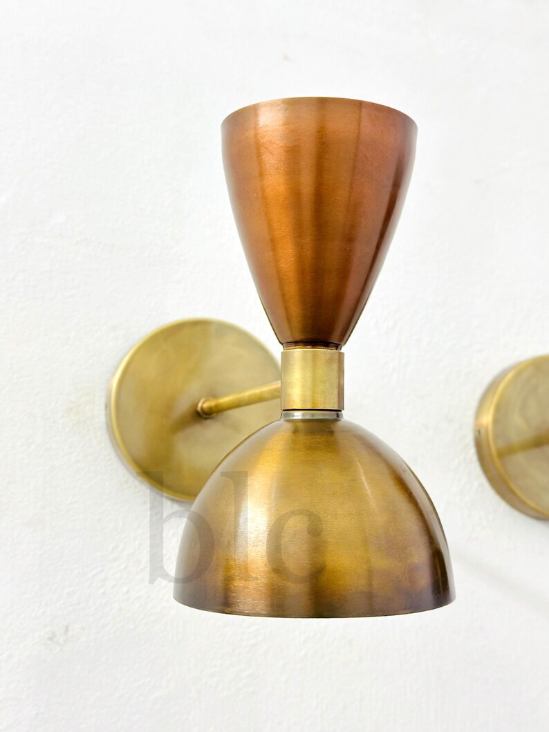May include: A pair of wall sconces with a mid-century modern design. The sconces feature a copper-colored conical shade and a brass-colored, dome-shaped base. The sconces are mounted on a white wall with brass-colored circular backplates.