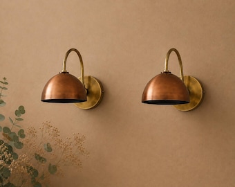 May include: Two wall-mounted lamps featuring dome-shaped copper shades and curved brass arms. Each lamp has a circular brass wall plate and a downward-facing opening. The design combines a metallic copper finish on the exterior with brass hardware.
