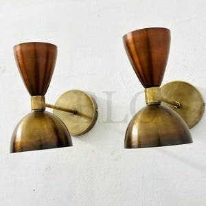 May include: Two vintage-style wall sconces. Each sconce features a copper-colored, cone-shaped shade atop a brass-colored, rounded base. The sconces are mounted on circular brass backplates, creating a warm, inviting aesthetic.