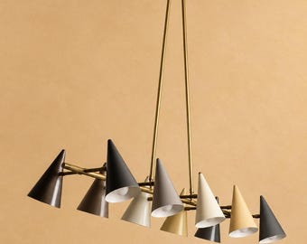 Peut inclure: Un lustre multi-lampes doté d'une tige centrale et d'un pavillon en laiton avec plusieurs bras s'étendant vers l'extérieur. Chaque bras supporte un abat-jour conique dans des tons variés de noir, gris, crème et beige, orientés vers le bas.