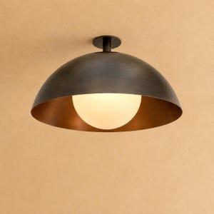 May include: A ceiling-mounted light fixture featuring a large, dark bronze-toned metal dome shade with a smooth exterior. Centered within the dome is a spherical, frosted white glass globe that emits a soft, diffused light from the interior.