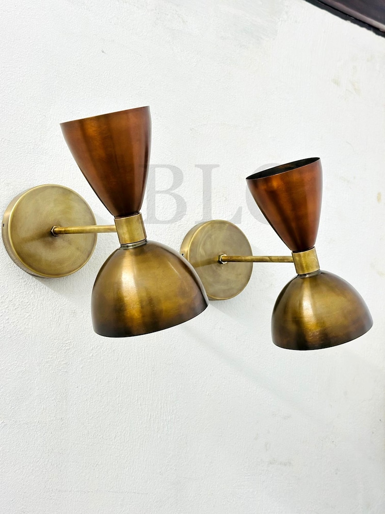 May include: Two wall-mounted sconces with a mid-century modern design. Each sconce features a brass-colored circular base, a slender arm, and a dual-tone shade. The upper portion of the shade is a copper color, while the lower is brass. The sconces are mounted on a white wall.