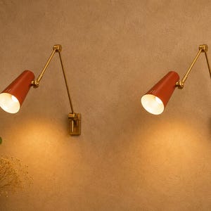 Könnte beinhalten: Ein Paar Wandleuchten mit verstellbaren Messingarmen und konischen orangefarbenen Schirmen. Jede Leuchte verfügt über eine runde Basisplatte und einen Gelenkarm, der nach außen ragt und das Licht aus dem orangefarbenen Metallkegel nach unten lenkt.