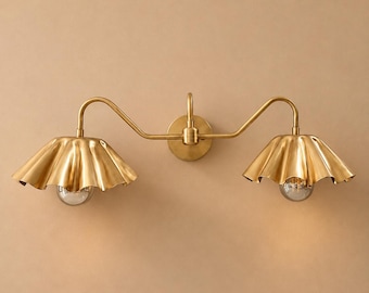 May include: A double-arm wall sconce featuring a polished brass finish. The fixture has a central circular mounting plate with two curved arms extending outward, each supporting a wide, ruffled, bell-shaped metal shade with a light bulb.