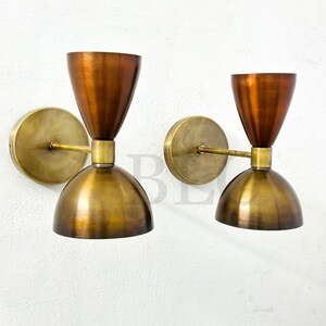 May include: Two mid-century modern wall sconces. Each sconce features a copper-colored conical shade above a brass-colored, dome-shaped base. The sconces are mounted on a round brass backplate with a brass arm. The fixtures are designed for wall mounting.