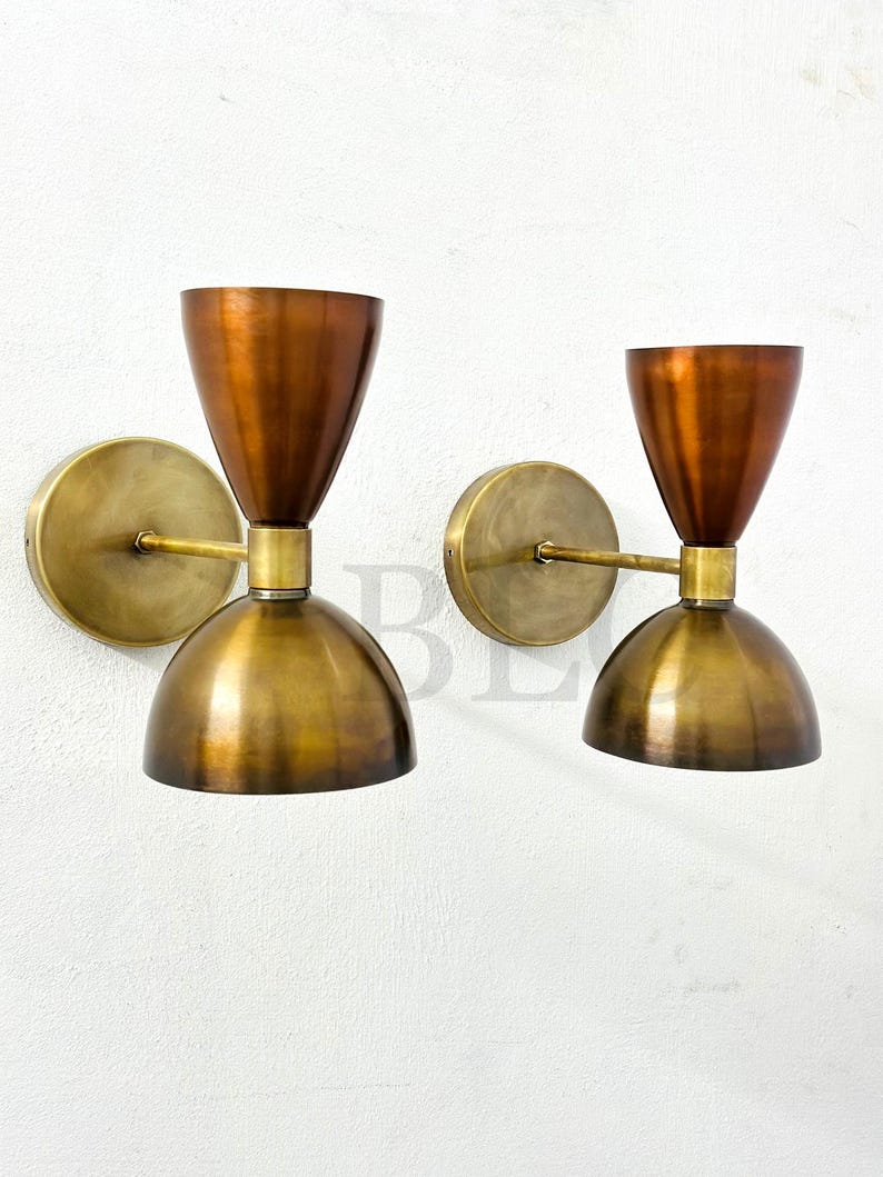 May include: Two mid-century modern wall sconces. Each sconce features a copper-colored conical shade above a brass-colored, dome-shaped base. The sconces are mounted on a round brass backplate with a brass arm. The fixtures are designed for wall mounting.