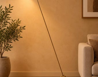 May include: A floor lamp with a long, thin brass-colored metal stand and a black, cone-shaped adjustable head. The lamp features a spherical base weight and a horizontal bar support, emitting a warm, bright light from the illuminated bulb.
