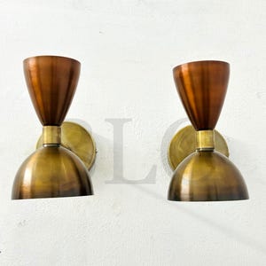 May include: Two vintage-style wall sconces. Each sconce features a copper-colored, cone-shaped shade atop a brass-colored, semi-spherical base. The sconces are mounted on a white wall, offering a warm, inviting glow.