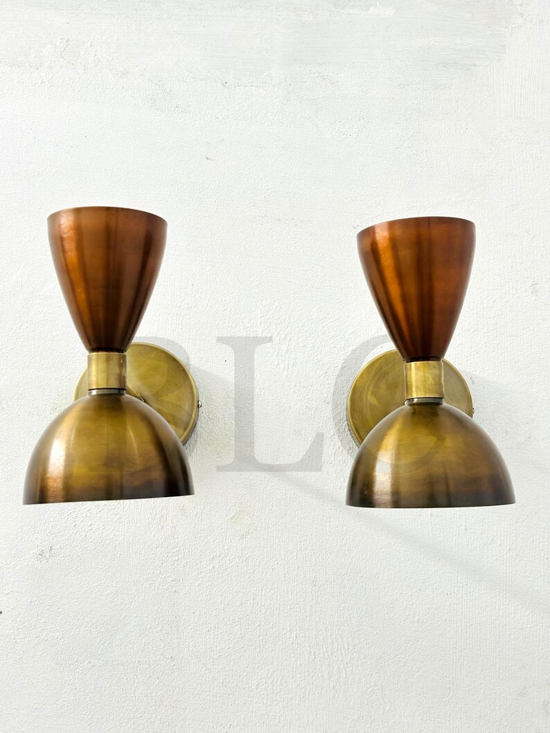 May include: Two vintage-style wall sconces. Each sconce features a copper-colored, cone-shaped shade atop a brass-colored, semi-spherical base. The sconces are mounted on a white wall, offering a warm, inviting glow.