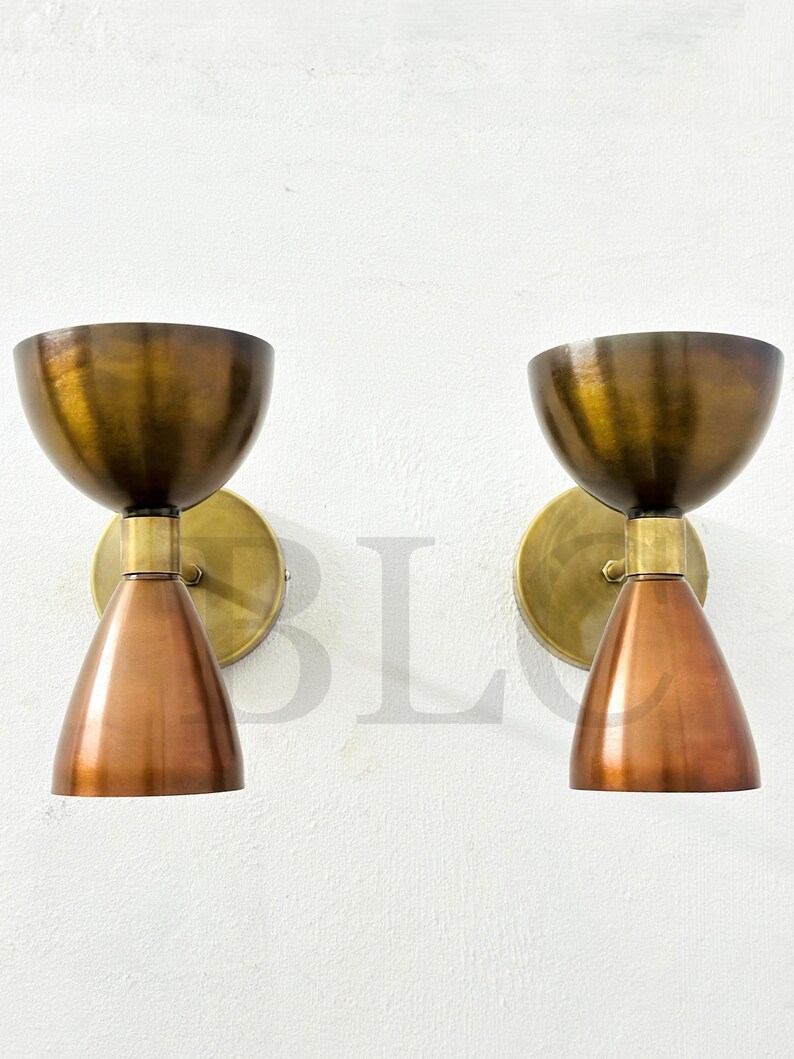 May include: Two vintage-style wall sconces. Each sconce features a copper-colored, cone-shaped shade and a brass-colored, bowl-shaped top. The sconces are mounted on round brass backplates, creating a warm, inviting aesthetic.