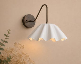 May include: A wall-mounted lamp featuring a dark bronze circular base and a curved arm supporting a white, wide, ruffled ceramic shade. The light fixture emits a warm glow from beneath the scalloped edge of the shade, creating a soft illumination.
