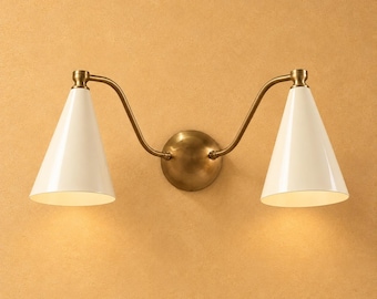 May include: A double wall sconce featuring a central circular brass mounting plate with two curved brass arms extending outward. Each arm supports a white conical shade pointing downward, creating a symmetrical lighting fixture design.