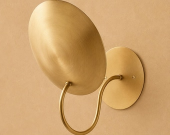 May include: A wall-mounted light fixture featuring a circular brass backplate and a curved arm that supports a large, shallow, bowl-shaped brass shade. The metal has a brushed, matte finish, and the design is composed of simple geometric shapes.