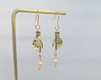 May include: Gold dangle earrings with a hand design and a pearl drop. The hand is pointing down.