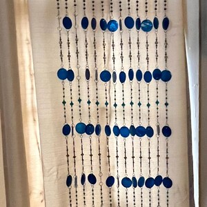 May include: A decorative wall hanging with strands of beads and blue, oval-shaped accents. The strands hang from a piece of driftwood. The beads are a mix of small, dark beads and larger, blue, oval-shaped beads.