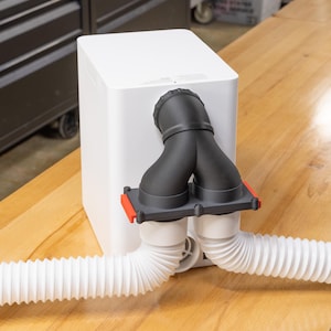 May include: A white rectangular device with a black Y-shaped nozzle and two white corrugated hoses. The nozzle is attached to a gray plate with red accents. The device sits on a light brown wooden surface.