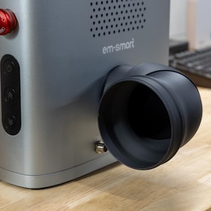 May include: A silver and black 3D printer with a black exhaust pipe and a control panel with buttons. The printer has the text "em-smart" on the front.