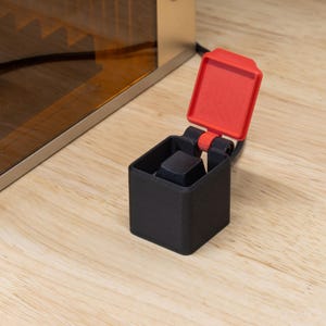 May include: A black, square-shaped 3D-printed object with a hinged red lid. The lid is open, revealing a black square inside. The object is on a light-colored wooden surface, near a metallic appliance.
