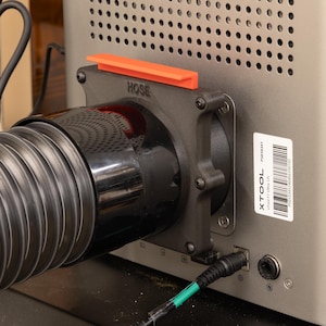 Könnte beinhalten: Ein schwarzer Kunststoff-Schlauchadapter mit einer orangefarbenen oberen Lasche und dem eingeprägten Wort HOSE auf der Vorderseite, befestigt an der Rückseite einer silbernen xTool F1 Ultra US Maschine. Der Adapter ist mit einem flexiblen Schlauch verbunden.