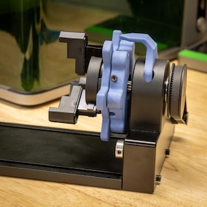 May include: A black and blue mechanical device with a complex design. The device features a series of gears, levers, and a blue, curved handle. The components are mounted on a black base, and the background is a light wood surface.