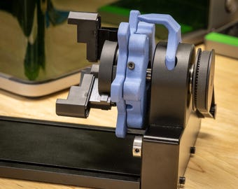 Op de afbeelding: Een zwart en blauw mechanisch apparaat met een complex ontwerp. Het heeft tandwielen, hendels en een blauwe, gebogen handgreep. De componenten zijn gemonteerd op een zwarte basis, met een lichte houten achtergrond.