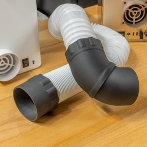 Op de afbeelding: Een flexibele witte gegolfde ventilatieslang met matzwarte kunststof aansluitingen aan beide uiteinden. Het ene uiteinde is een rechte cilindrische connector, terwijl het andere uiteinde een gebogen elleboogstuk heeft voor gerichte luchtstroom.