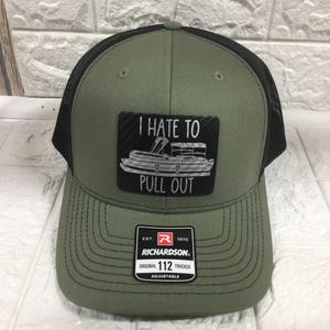 May include: Olive green trucker hat with a black mesh back. The front features a patch with the text "I HATE TO PULL OUT" and a pontoon boat graphic. The hat has a curved brim and an adjustable strap.
