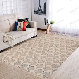 Puede incluir: Alfombra beige con un patrón de plumas grises repetidas. La alfombra tiene un borde con flecos y está colocada sobre un suelo de madera. Un sofá crema y cojines decorativos están en el fondo, junto con una foto enmarcada de la Torre Eiffel.