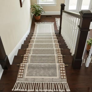 Peut inclure: Un tapis de couloir pour escalier avec un motif géométrique beige et marron. Le tapis est orné de franges en bas et est installé sur des marches en bois foncé. Un élément décoratif pour l'intérieur.