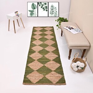 Puede incluir: Una alfombra larga y rectangular con un patrón de diamantes verdes y beige. La alfombra está hecha de fibras naturales y está colocada sobre un suelo blanco. La alfombra está frente a un banco y una pequeña mesa auxiliar. Tres estampados botánicos enmarcados cuelgan de la pared.