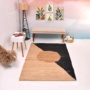 Op de afbeelding: Een geometrisch jute vloerkleed met een beige en zwart ontwerp. Het vloerkleed heeft een grote beige cirkel en een driehoek van beige en zwart. Het vloerkleed ligt op een witte vloer, met een bank en kunstwerken op de achtergrond.