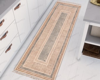 Puede incluir: Un tapete corredor beige y marrón con diseños geométricos en blanco y negro. El tapete tiene flecos en los extremos y está colocado sobre un suelo blanco.
