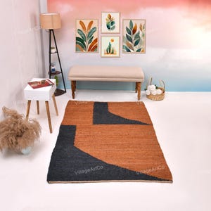May include: A rectangular rug with a geometric design. The rug has a burnt orange background with two large, asymmetrical dark grey shapes. The rug is on a white floor, with a bench, artwork, and a lamp in the background.