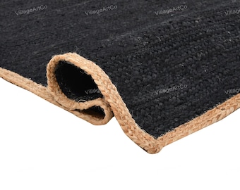 May include: A rolled-up black jute rug with a natural tan border. The rug's texture is visible, and the border is tightly woven. The words "VillageArtCo" are printed on the rug.