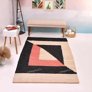 May include: A rectangular jute rug with a geometric design. The rug features a black square with a coral pink and off-white triangle in the center. The border is off-white. The rug is on a white floor.