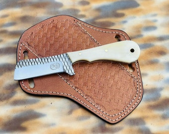 Puede incluir: Un pequeño cuchillo de acero inoxidable con mango de hueso y hoja texturizada. El cuchillo está en una funda de cuero marrón con un patrón tejido.
