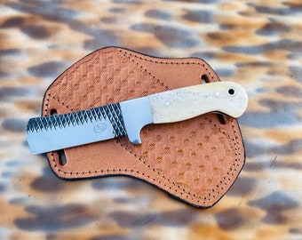 Puede incluir: Un cuchillo artesanal con mango color crema y hoja dentada, sobre una funda de cuero marrón. La funda tiene un patrón texturizado y bordes cosidos. La hoja del cuchillo tiene las iniciales "KJ" grabadas.