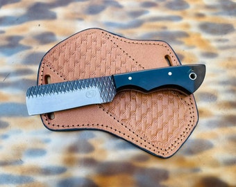 Puede incluir: Un cuchillo con mango negro y hoja texturizada, en una funda de cuero. El cuchillo tiene un pequeño agujero en el mango y un patrón decorativo en la hoja.