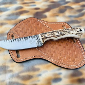 May include: A fixed-blade knife with a textured steel blade featuring a circular logo, a handle made of natural antler with three metal pins, and a lanyard hole, resting on a tan leather sheath with a basket-weave pattern and white stitching.