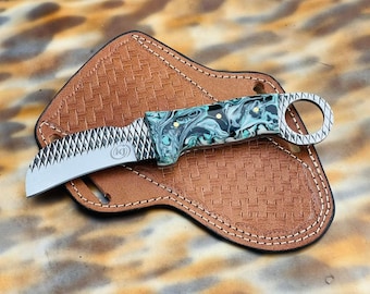 May include: A curved, stainless steel knife with a black and green marbled handle. The knife has a serrated edge and a small ring on the handle. The knife is resting on a brown leather sheath.