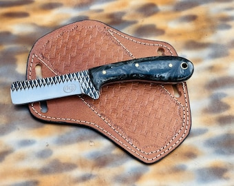 Puede incluir: Un cuchillo de hoja fija con una hoja de metal texturizada tipo escofina que presenta un logotipo circular con las letras KJ, un mango de resina veteada en blanco y negro con tres pasadores de latón y un orificio para cordón, sobre una funda.