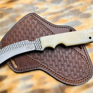 May include: A curved, stainless steel knife with a white bone handle and a file pattern on the blade. The knife is in a brown leather sheath with a woven pattern.